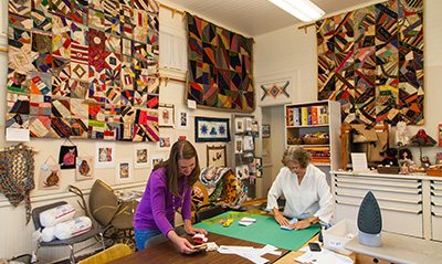 Two women cut and measure fabric with quilts mounted on the walls around them