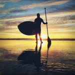 Silhouette of a person holding an oar and a paddle board in front of the sunset