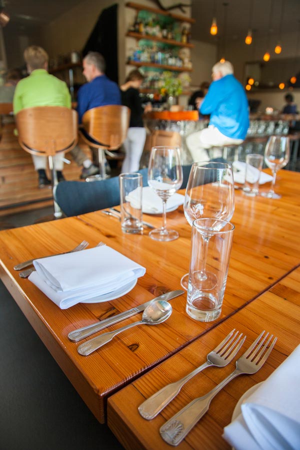 Wooden table in restaurant set with silverware and wine glasses, people sitting on bar stools in background