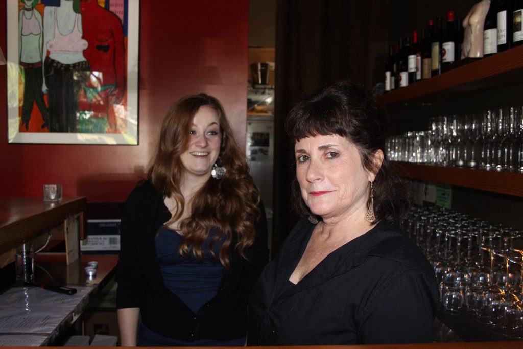 Two women stand behind the bar with wall of wine glasses behind them