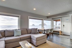 Gray living room with long couch, dining table and windows looking out on the ocean