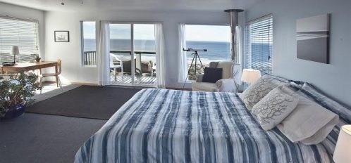 Small bedroom with blue bedspread and telescope next to window that looks out on the beach