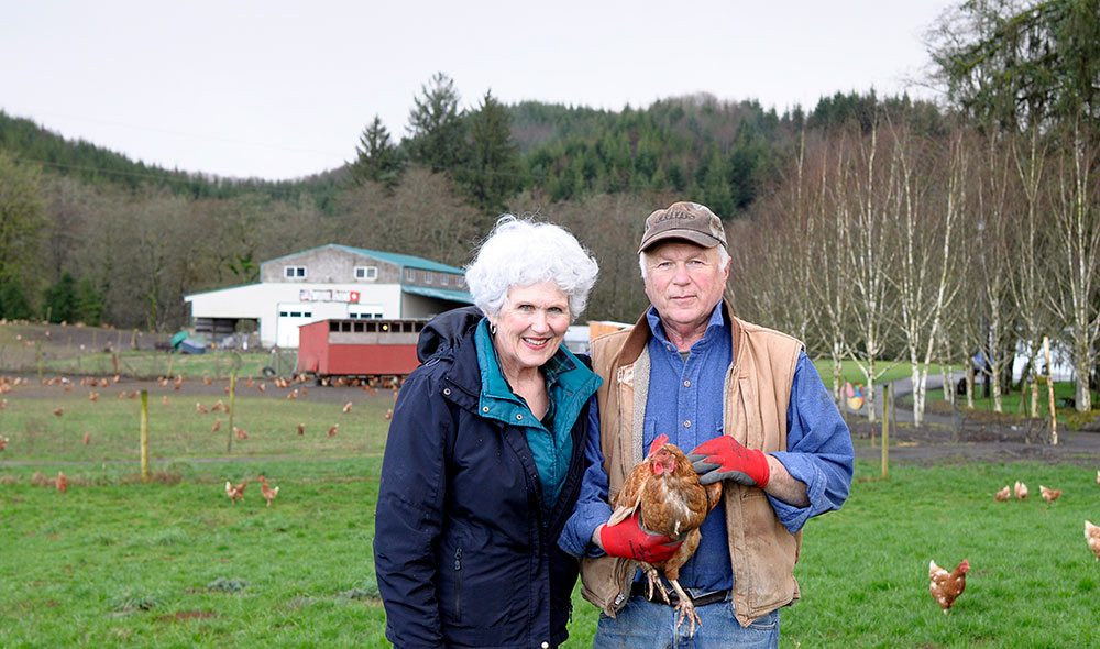 Man and woman stand on farm holding a small brown hen with more chickens in background